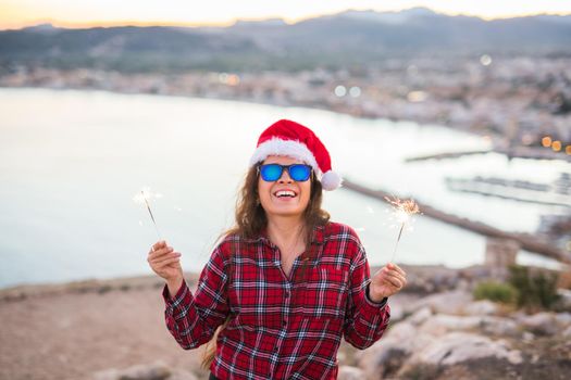 Holiday, Christmas and people concept - Young happy woman in Santa hat over beautiful landscape with fireworks and sparklers.