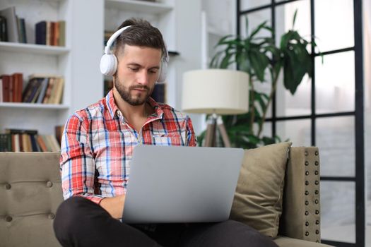 Concentrated young freelancer businessman sitting on sofa with laptop, working remotely online at home