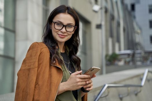 Business lady. Portrait of a young beautiful happy caucasian woman holding her smartphone and smiling at camera while standing on the city street. Urban lifestyle concept