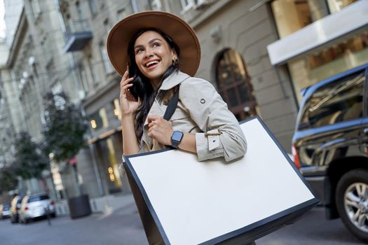 Shopping makes us happy. Young beautiful excited woman in hat with shopping bag talking on smartphone, looking aside and smiling while walking on the city streets. Fashion, people lifestyle concept