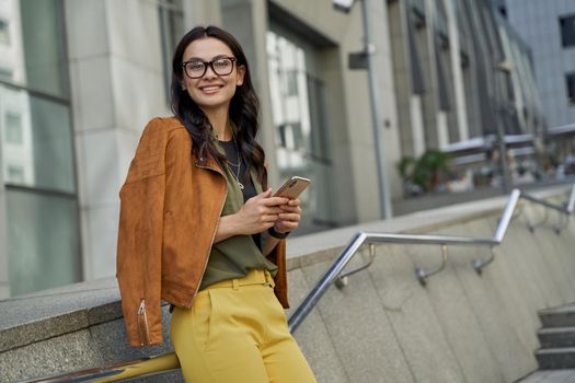 Texting sms to friend. Young beautiful and fashionable caucasian woman holding her smartphone, looking at camera and smiling while standing on the city street, business lady outdoors. People lifestyle