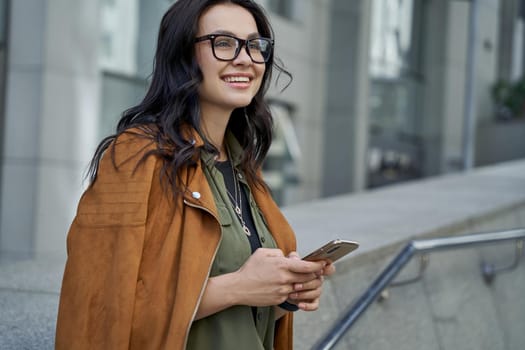 Walking on the city streets. Young beautiful and fashionable woman wearing eyeglasses using her smartphone, chatting with friend and smiling while standing against blurred urban background