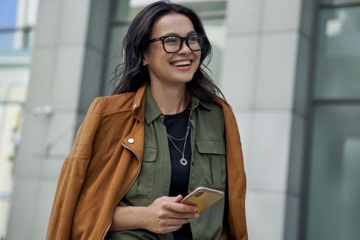 Being outside on a beautiful day. Young happy fashionable woman wearing eyeglasses walking on the city street, using her smartphone and smiling while standing against blurred urban background