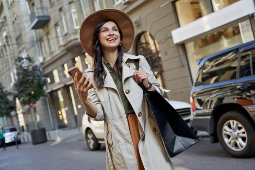 Good feelings after shopping. Young beautiful stylish woman in hat with shopping bag holding her smartphone, looking aside and smiling while walking on the city streets. Fashion, people lifestyle