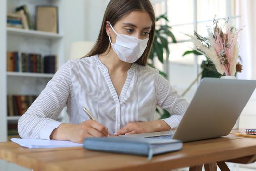 Young businesswoman in a medical protective mask works from home at the laptop during self-isolation and quarantine