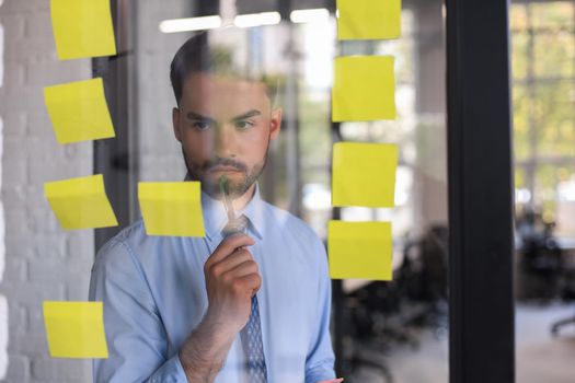 Young modern business man using adhesive notes while standing behind the glass wall in the office