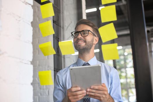 Young modern business man using adhesive notes while standing behind the glass wall in the office