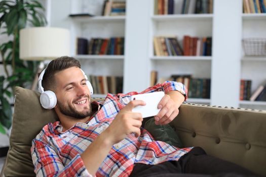Attractive young man relaxing on a couch at home, listening music and playing on smartphone