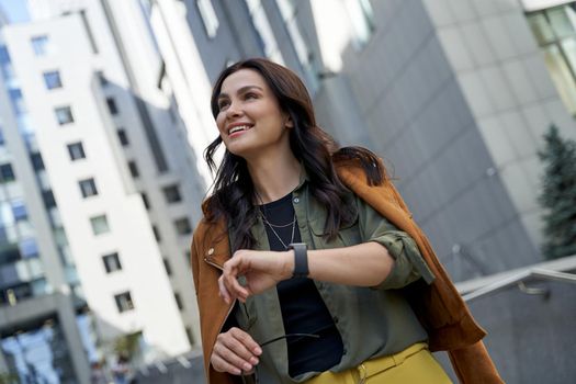 Portrait of a young beautiful and happy business woman checking time on the watch on her hand while standing on the city street, looking aside and smiling. Urban lifestyle concept