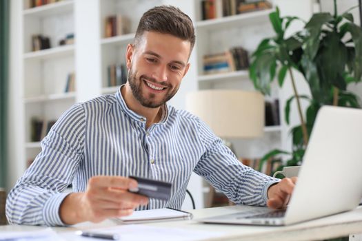 Smiling man sitting in office and pays by credit card with his laptop