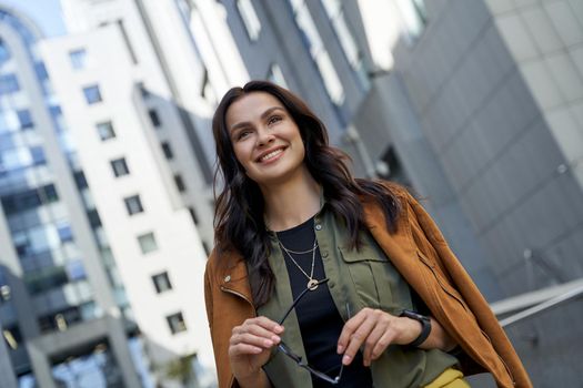 Portrait of a young beautiful and happy business lady with eyeglasses in hand standing on the city street, looking aside and smiling. Urban lifestyle concept, business people