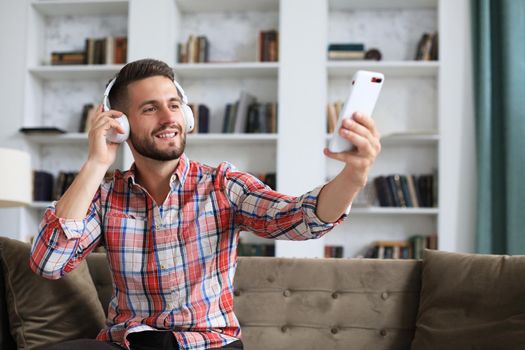 Attractive man taking selfie photo or self video with mobile phone at home sitting on couch
