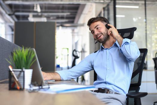 Smiling male call-center operator with headphones sitting at modern office, consulting online information in a laptop, looking up information in a file in order to be of assistance to the client