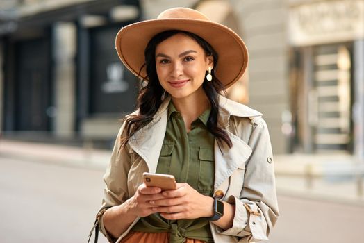 Portrait of a young beautiful stylish woman wearing autumn coat and hat holding her smartphone, looking at camera and smiling while standing on city street. Fashion, shopping, people lifestyle