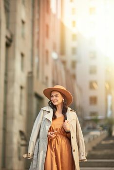 Vertical shot of a young beautiful and stylish caucasian woman wearing long romantic dress and hat looking aside and thinking about something while standing on the city street, walking outdoors