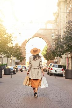 Rear view of a stylish woman wearing grey coat and hat with shopping bags walking city streets, full length. Shopaholic, fashion, people lifestyle concept