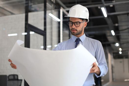 Confident young architect in hardhat holding a blueprint and looking at area for work