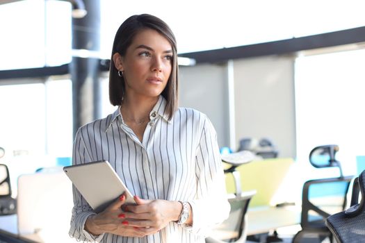 Portrait of young businesswoman holding touch pad while standing in modern office space interior