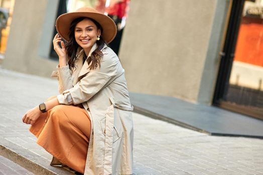 Enjoying nice warm day outdoors. Young attractive happy woman wearing autumn coat and hat looking at camera and smiling while walking city streets. People lifestyle concept