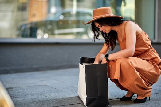 Young caucasian woman wearing long romantic dress and hat, looking in her shopping bag while squatting down on the city street. People lifestyle and fashion concept