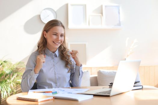 Happy entrepreneur woman sit at desk reading good news and express joy rising hands up