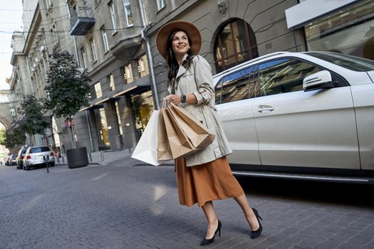 Shopping enjoyment. Full length of a young stylish business woman with paper shopping bags looking aside and smiling while standing on the city street against her white luxury car. People lifestyle