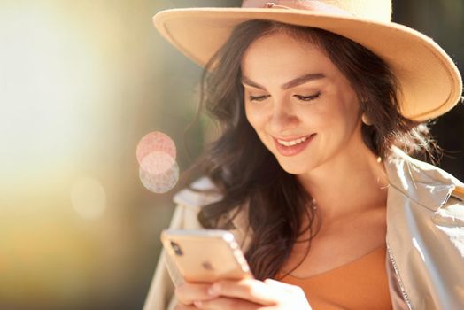 Texting sms. Portrait of a young beautiful and happy caucasian woman wearing hat using smartphone and smiling while standing on the city street on a sunny day