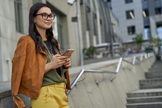 Side view of a young beautiful and fashionable caucasian woman using her smartphone and smiling while spending time outdoors, standing on the city street. People lifestyle