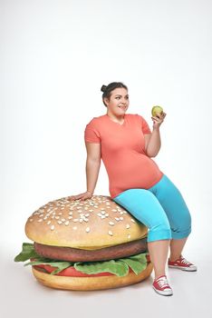 Funny picture of brunette, bearded, plump woman on white background. Woman wearing sportswear. She laid crouched on a huge sandwich