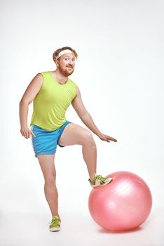 Funny picture of red haired, bearded, plump man on white background. Man wearing sportswear. Man put his leg to a fitness ball