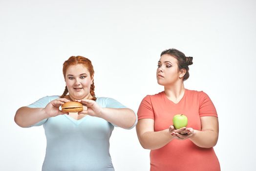 Funny picture of amusing chubby women on white background. One woman holding a sandwich, the other holding an apple.