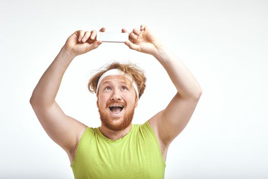 Funny picture of red haired, bearded, plump man on white background. Man wearing sportswear. Man smiling and taking a selfie