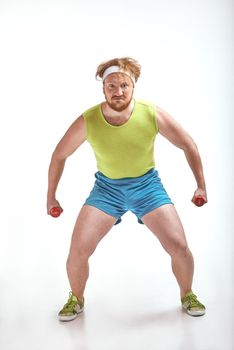 Funny picture of red haired, bearded, plump man on white background. Man wearing sportswear. Man holding the dumbbells