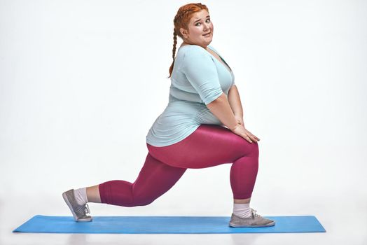 Funny picture of amusing, red haired, chubby woman on white background. Woman trains on the mat.