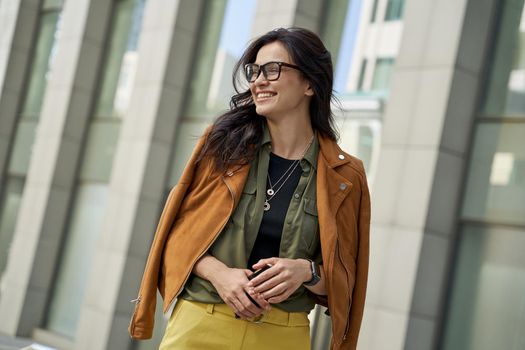 Portrait of a young happy woman holding smartphone, looking aside and smiling while walking on the city street on autumn day, stylish lady standing against blurred urban background. People lifestyle