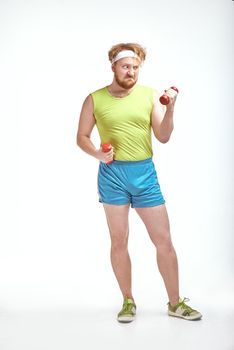 Funny picture of red haired, bearded, plump man on white background. Man wearing sportswear. Man holding the dumbbells