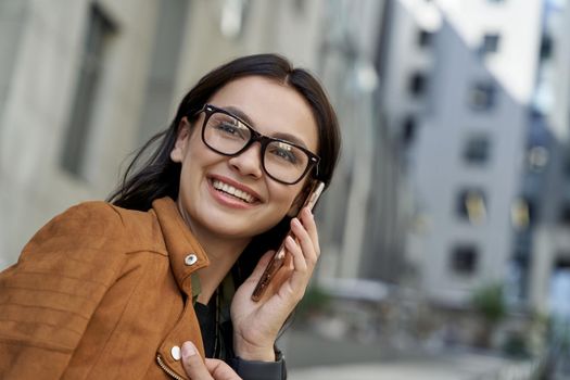 Close up portrait of a young beautiful woman wearing eyeglasses talking on smartphone with friend, looking aside and smiling while standing on the city street. Urban lifestyle concept, people