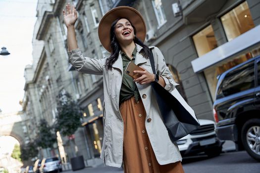 Shopping addiction. Young beautiful stylish woman in hat with shopping bag holding her smartphone and waving to friend while standing on the city street. Fashion, people lifestyle, retail therapy