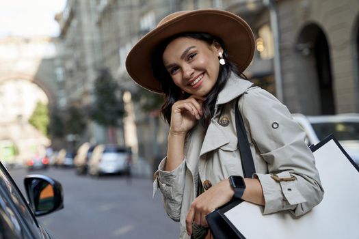 Shopping makes me feel good. Portrait of a young beautiful and happy caucasian woman in hat with shopping bag looking at camera and smiling while standing on the city street. Fashion, people lifestyle