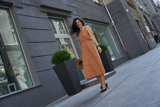 Fashion girl. Full length of a young beautiful and stylish caucasian woman wearing long romantic dress and high heel shoes holding hat, looking at camera and smiling while standing on the city street