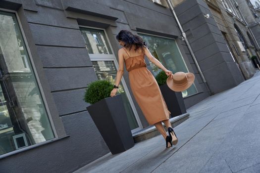 Full length shot of a young beautiful and stylish caucasian woman wearing long romantic dress holding hat feeling happy while walking outdoors, standing on the city street. Fashion, people lifestyle