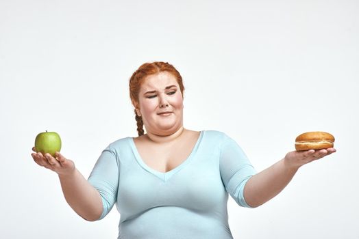 Funny picture of amusing, red haired, chubby woman on white background. Woman holding apple and sandwich. She is looking at a sandwich