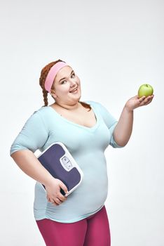 Funny picture of amusing, red haired, chubby woman on white background. Woman holding an apple and scales.