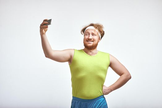 Funny picture of red haired, bearded, plump man on white background. Man wearing sportswear. Man smiling and taking a selfie