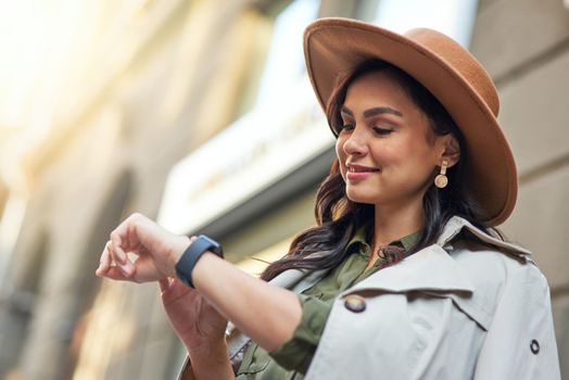 Young smiling stylish woman wearing grey coat and hat checking the time while standing on the city street, enjoying walking on warm autumn day. Beautiful people concept