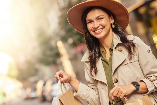 Visiting fashion boutique. Portrait of a young excited woman wearing grey coat and hat carrying shopping bags, standing on city street and smiling. Shopaholic, fashion, people lifestyle concept