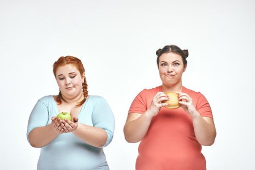 Funny picture of amusing chubby women on white background. One woman holding a sandwich, the other holding an apple.