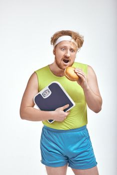 Funny picture of red haired, bearded, plump man on white background. Man holding a sandwich and scales