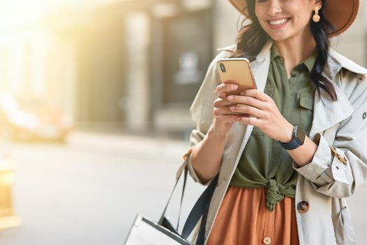 Cropped shot of a young beautiful happy woman wearing autumn coat and hat using her smartphone, chatting with friend and smiling while standing on city street. Fashion, shopping, people lifestyle