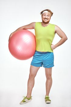Funny picture of red haired, bearded, plump man on white background. Man wearing sportswear. Man holding fitness ball